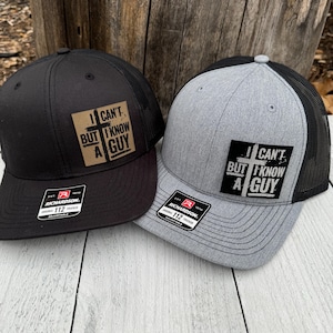 May include: Two trucker hats, one black and one gray, with a mesh back and a curved brim. Each hat has a patch that reads "I CAN'T BUT I KNOW A GUY". The hats are adjustable and have a Richardson 112 tag.