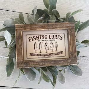 May include: A framed sign with the text "FISHING LURES" and illustrations of fishing lures. The sign is in a rustic wooden frame and surrounded by green and gray foliage. The text "BUILT FOR THE WATER - MADE TO LAST" is also visible.