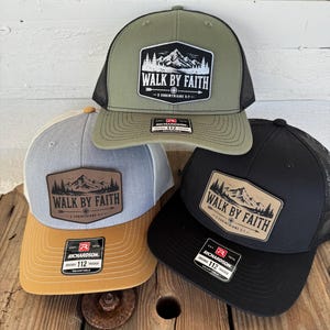 May include: Four trucker hats in olive green, gray, black, and tan. Each hat features a patch with a mountain scene and the text "WALK BY FAITH - 2 CORINTHIANS 5:7". The hats have a mesh back and adjustable fit.