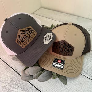 May include: Two trucker hats with the text "Faith Can Move Mountains." One hat is gray with a white mesh back, the other is tan with a brown mesh back. Both have a mountain graphic on a brown patch.