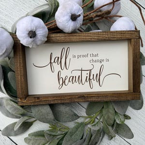 May include: A wooden framed sign with the words "fall is proof that change is beautiful" in brown script. The sign is surrounded by faux white pumpkins and green foliage, creating a fall-themed decorative piece.