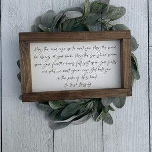 May include: A rectangular wooden sign with a white background and black script. The text is an Irish blessing. The sign is surrounded by a wreath of green leaves, set against a white wood-paneled background.