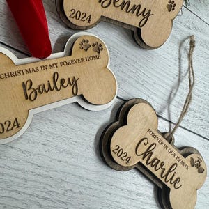 May include: Three wooden dog bone ornaments with the names "Benny", "Bailey", and "Charlie" engraved on them. The ornaments are decorated with paw prints and the year "2024".