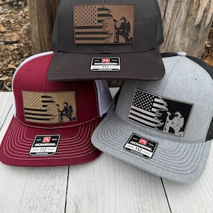 May include: Three trucker hats with a patriotic theme. The hats are black, maroon, and gray, each featuring a leather patch with a distressed American flag and a firefighter silhouette. The hats have a mesh back and a curved brim.