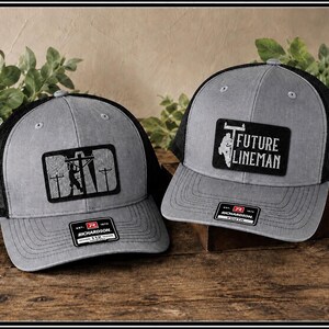 Peut inclure: Deux casquettes de camionneur grises avec des dos en filet noirs. L'une des casquettes présente un écusson avec une silhouette de ligneur et le mot "DAD". L'autre casquette porte un écusson avec l'inscription "FUTURE LINEMAN". Les deux casquettes ont une étiquette Richardson 112.