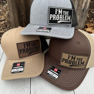 May include: Three trucker hats in gray, tan, and brown with the text "I'M THE PROBLEM" on a rectangular patch. Each hat has a curved brim and a mesh back. The hats are adjustable and have a Richardson 112 Trucker tag.