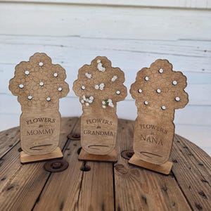 May include: Three wooden flower-shaped decorations. Each has a vase design with the words "Flowers for Mommy", "Flowers for Grandma", and "Flowers for Nana". The flowers are laser-cut with small white accents, and sit on a wooden base.
