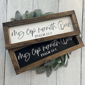 May include: Two rectangular wooden signs with the text "My Cup runneth Over, Psalm 23:5" in white script. One sign has a white background, the other is black. The signs are framed in dark brown wood and decorated with green foliage.