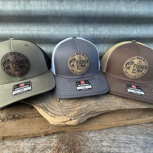 May include: Three trucker hats in olive green, gray, and brown, each with a mesh back and a curved brim. The hats feature a circular patch with the text "JESUS MADE ME A FISHER OF MAN" and a fish graphic. The hats are displayed on a wooden surface.