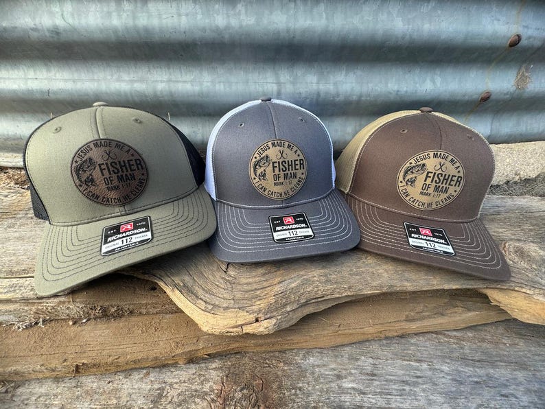 May include: Three trucker hats in olive green, gray, and brown, each with a mesh back and a curved brim. The hats feature a circular patch with the text "JESUS MADE ME A FISHER OF MAN" and a fish graphic. The hats are displayed on a wooden surface.