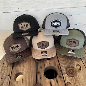 May include: Six trucker hats in various colors, including black, gray, brown, and olive green. Each hat features a patch with the words "DAD" or "PAPA" in a bold font. The hats are displayed on a wooden spool.