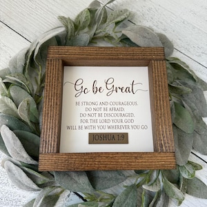 May include: A wooden framed sign with the text "Go be Great" and a Bible verse from Joshua 1:9. The sign is surrounded by a wreath of green and grey leaves.