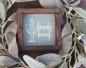 May include: A rustic wooden sign with a light gray background featuring a white silhouette of a cactus and the text "Howdy Honey".