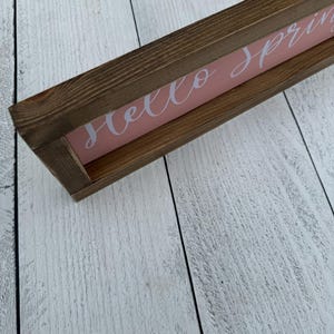 May include: A rectangular wooden sign with a brown frame and a pink background. The sign reads "Hello spring" in white cursive lettering. The sign is placed on a white wooden surface.