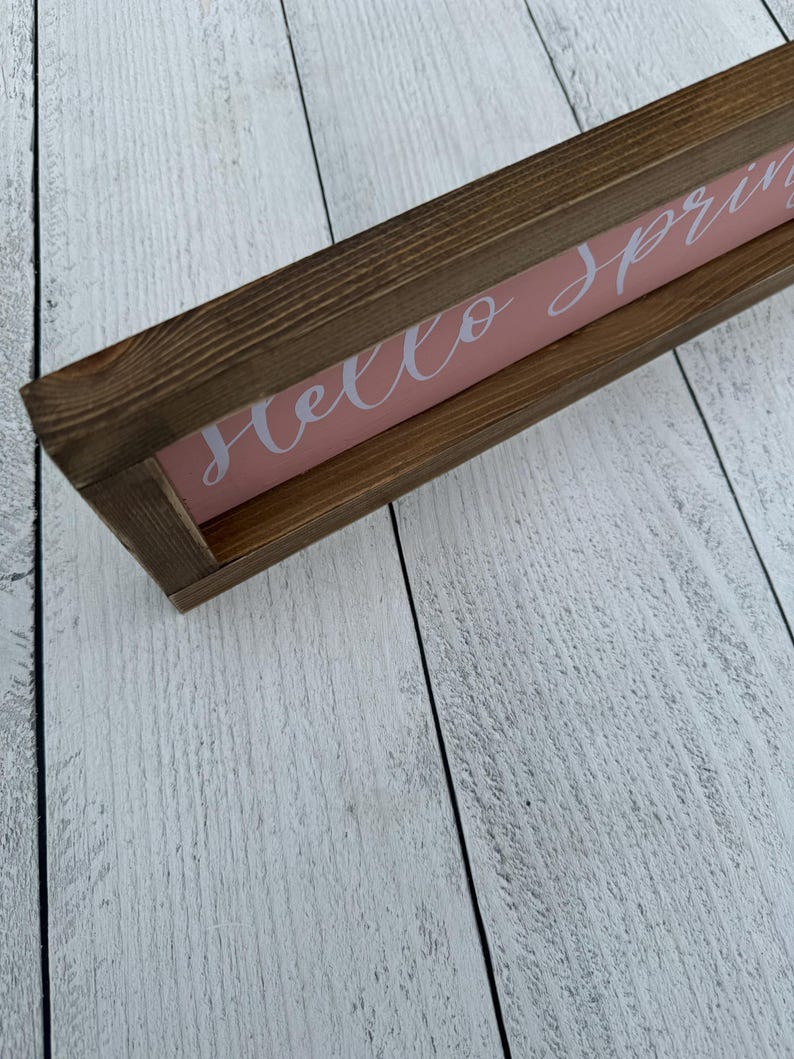 May include: A rectangular wooden sign with a brown frame and a pink background. The sign reads "Hello spring" in white cursive lettering. The sign is placed on a white wooden surface.