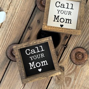 K&ouml;nnte beinhalten: Zwei kleine, quadratische Holzschilder mit dem Text "Call Your Mom" in wei&szlig;en Buchstaben und einem Herzsymbol. Ein Schild hat einen schwarzen Hintergrund, das andere einen cremefarbenen Hintergrund. Die Schilder werden auf einer rustikalen Holzoberfl&auml;che angezeigt.