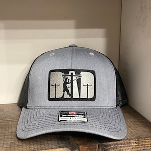 Peut inclure: Un chapeau de camionneur gris et noir avec une silhouette noire d'un lineman travaillant sur une ligne électrique. Le chapeau a un dos en maille gris et une visière noire. Le devant du chapeau a un patch blanc avec le mot "DAD" en lettres noires. Le chapeau a une étiquette noir et blanc avec le texte "Richardson 112" sur le devant.