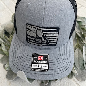 May include: A grey and black trucker hat with a black and white American flag design featuring a welder. The hat has a red, white and black label that reads "EST. 1970 RICHARDSON ORIGINAL 112 TRUCKER ADJUSTABLE".