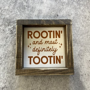 May include: A wooden sign with a white background and brown text that reads "ROOTIN' and most definitely TOOTIN'".