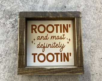 Puede incluir: Un letrero de madera con fondo blanco y texto marrón que dice "ROOTIN' and most definitely TOOTIN'".
