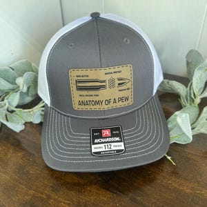 May include: A gray and white trucker hat with a brown patch that says "Anatomy of a Pew" and depicts a bullet with labels for the different parts. The hat is adjustable and has a Richardson 112 logo.