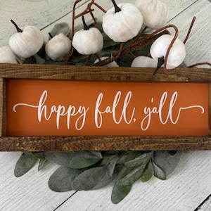 May include: A rectangular wooden sign with an orange background and the words "happy fall, y'all" in white script. The sign is surrounded by white pumpkins, greenery, and brown stems, creating a fall-themed decoration.