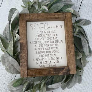 May include: A framed wooden sign with the text "The Ten Commandments" in elegant script. The sign lists the commandments in a clear, readable font. The frame is a dark brown color, and the sign is surrounded by a wreath of green leaves.