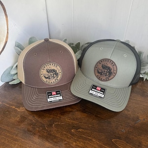 May include: Two trucker hats in brown and green with a black mesh back. The hats feature a brown leather patch with a fish and the text "Weekend Hooker" on the front.