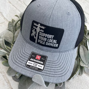 May include: A grey and black trucker hat with a black leather patch that says "Support Your Local Pole Dancer". The hat is adjustable and has the Richardson 112 logo on the side.