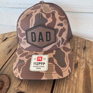 May include: A camouflage print baseball cap with a brown mesh back. The front of the cap features a black hexagon patch with the word "DAD" in bold letters. A white rectangular tag with text is visible on the front.