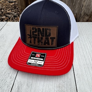 May include: A Richardson Original 112 Trucker hat. It has a red peak, navy blue crown, white mesh back, and a brown leather patch. The patch features the text "2ND THAT" with rifle illustrations. The hat has an adjustable strap.