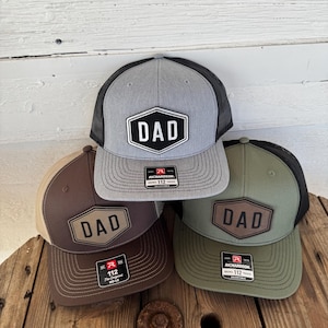 May include: Four trucker hats with the word "DAD" on a patch. The hats are in gray, brown, and olive green with black mesh backs. Each hat has a Richardson 112 tag.