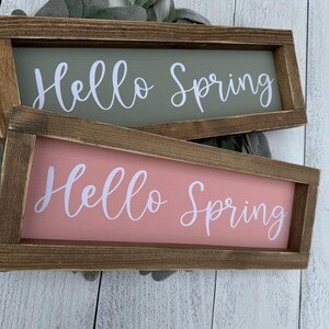 May include: Two rectangular wooden signs with the text "Hello Spring" in white script. One sign is sage green, the other is coral. The signs are framed in brown wood and rest on a white wooden surface.
