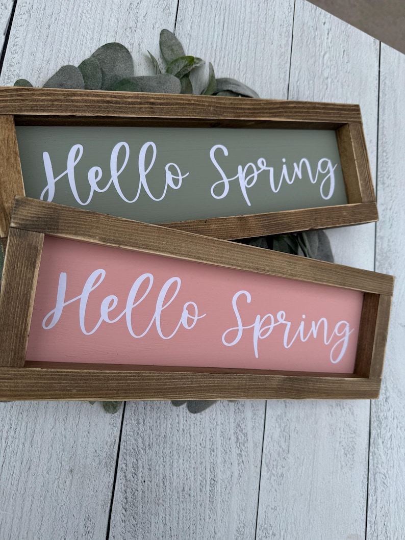 May include: Two rectangular wooden signs with the text "Hello Spring" in white script. One sign is sage green, the other is coral. The signs are framed in brown wood and rest on a white wooden surface.