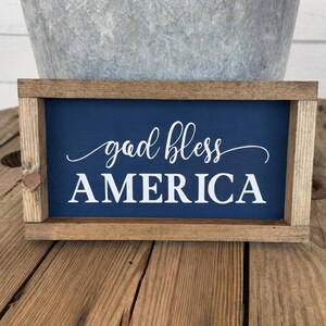 Puede incluir: Cartel enmarcado de madera con fondo azul marino y las palabras "god bless AMERICA" en escritura blanca. El marco es de madera marr&oacute;n claro. Un cubo de metal est&aacute; en el fondo.