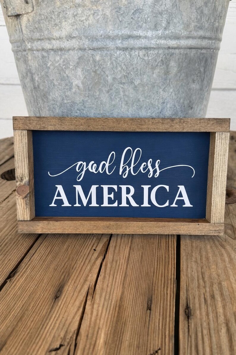 Puede incluir: Cartel enmarcado de madera con fondo azul marino y las palabras "god bless AMERICA" en escritura blanca. El marco es de madera marr&oacute;n claro. Un cubo de metal est&aacute; en el fondo.