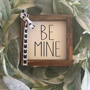 May include: A wooden sign with the text "BE MINE" in black lettering on a white background. The sign is framed in a dark brown wood frame and has a black and white ribbon tied around it.