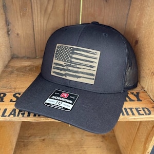 May include: Black mesh trucker hat with a brown distressed American flag patch featuring a silhouette of rifles. The hat has a black Richardson 112 adjustable label.