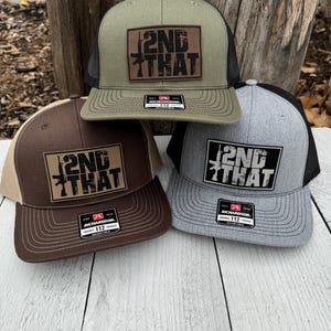 May include: Three trucker hats in olive green, brown, and gray, each featuring a rectangular patch with the text "2ND THAT" and a rifle graphic. The hats have mesh backs and curved visors.