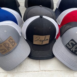 May include: A collection of eight trucker hats with various colors and designs. Some hats feature a brown leather patch with embroidered text, including "SRB" and "Jayden".