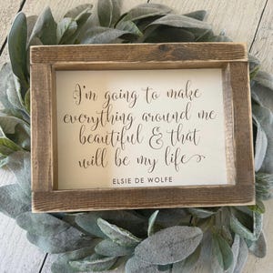 May include: A wooden framed sign with the quote "I'm going to make everything around me beautiful & that will be my life" by Elsie De Wolfe. The sign is surrounded by a green leafy wreath.