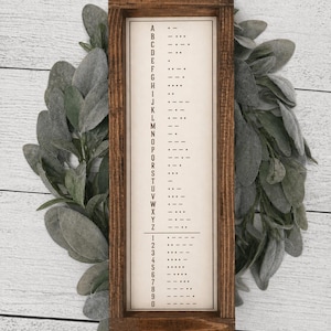 May include: A rectangular wooden sign with a brown frame displays a Morse code alphabet and numbers. The sign's background is off-white, with black dots and dashes representing each letter and number. The sign is surrounded by green foliage.