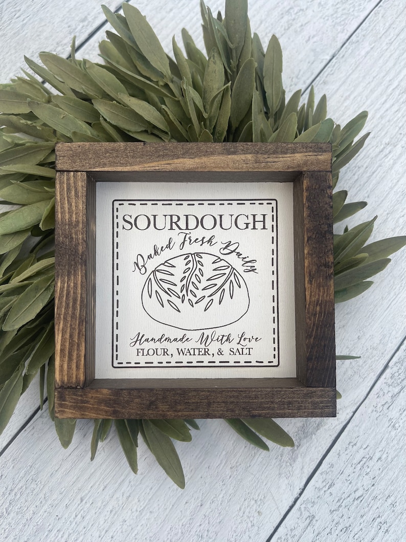 Pu&ograve; includere: Un cartello in legno rustico con sfondo bianco e testo nero che recita "SOURDOUGH Baked Fresh Daily Handmade With Love FLOUR, WATER, & SALT". Il cartello &egrave; incorniciato in una cornice di legno marrone scuro.