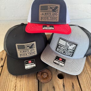 May include: A selection of Richardson 112 trucker hats in navy, black, and grey. Each hat displays a patch with an American flag and the text "ALWAYS LOVE YOUR COUNTRY NEVER TRUST YOUR GOVERNMENT". The hats have mesh backs and curved peaks.