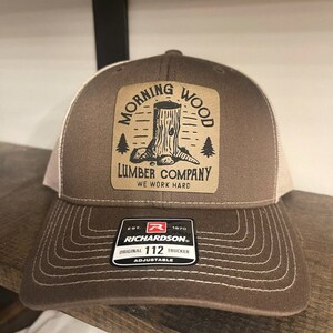 May include: Brown and white trucker hat with a brown patch that says "Morning Wood Lumber Company We Work Hard". The patch features a cartoon drawing of a tree stump. The hat also has a black and white label that says "Richardson Original 112 Trucker Adjustable".