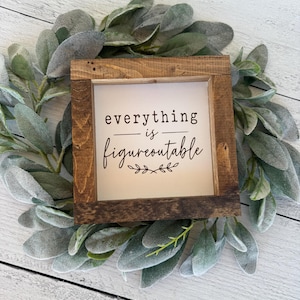 May include: A square wooden sign with a dark brown stained frame and a white center panel. The text "everything is figureoutable" is printed in black, with a small decorative leaf flourish underneath the lettering, centered on the sign.