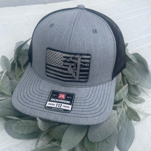 May include: A gray and black trucker hat with an American flag patch featuring a silhouette of a lineman working on a power line. The hat has a black mesh back and a red label that reads "Richardson 112 Trucker Adjustable".