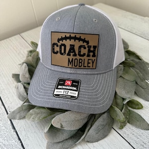 May include: A gray and white trucker hat with the text "COACH MOBLEY" on a brown patch. The hat has a curved brim and an adjustable strap. The hat is a Richardson 112 Trucker.