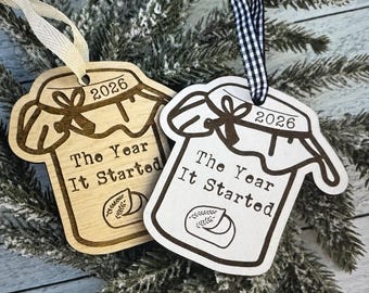 May include: Two wooden jar-shaped ornaments featuring the engraved text 2026 and The Year It Started, with a small illustration of a sourdough loaf. One ornament has a cream ribbon, the other a black and white gingham ribbon for hanging.
