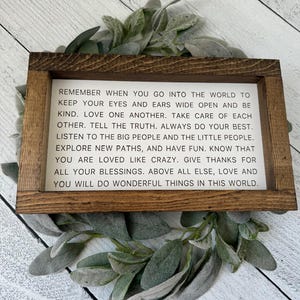 May include: A rectangular wooden sign with a white background and black text. The sign is surrounded by a green and gray leafy garland. The text on the sign reads: "REMEMBER WHEN YOU GO INTO THE WORLD..."